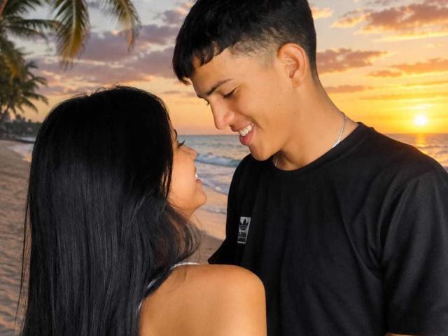 NatashaxxxRyan with long black hair and small tits, smiling at a young dude with a short haircut and black shirt, standing on a beach at sunset.