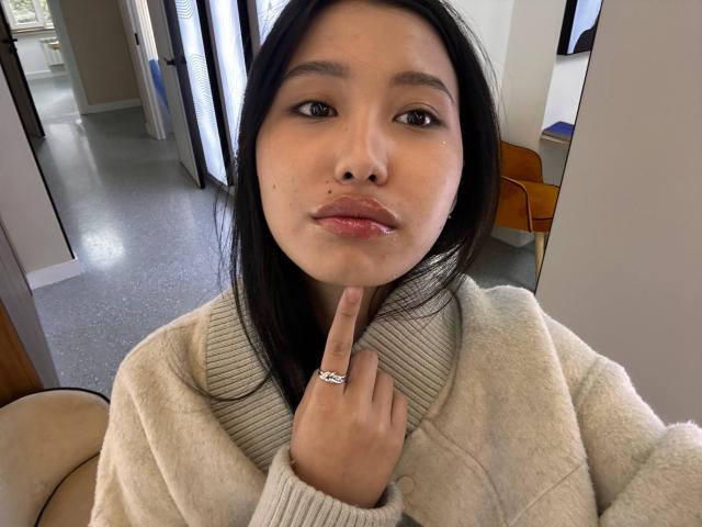 LeonilaSeewald with glossy pink lips, black straight hair, light brown skin, wearing a fluffy beige sweater, finger on chin with a silver ring, in a bright indoor setting.