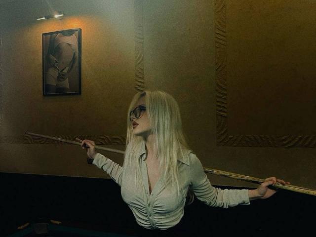 JerleneLeoni with blonde hair, glasses, and a white button-up shirt, holding a pool cue, leaning forward, showing her big tits and small ass. Dark room, framed picture.