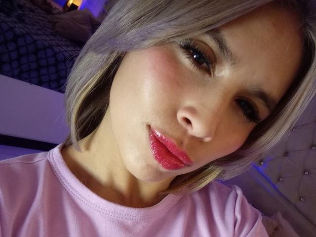 MacarenaJonesFg with shiny pink lips, blushed cheeks, and glossy eyebrows, wearing a pink shirt, winking at the camera, blonde hair, and cute face.