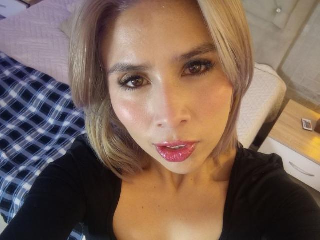 MacarenaJonesFg has shiny brown eyes, glossy pink lips, and light brown hair in a bob. She's wearing a black top, looking at the camera with a slight pout.
