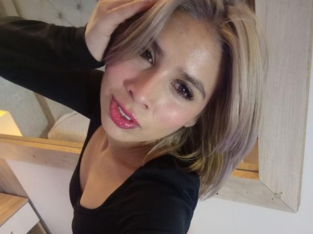 MacarenaJonesFg with shiny pink lips, black top, and blonde hair, hand in hair, smiling at camera, showing ass in the background, soft light, warm skin, relaxed pose.