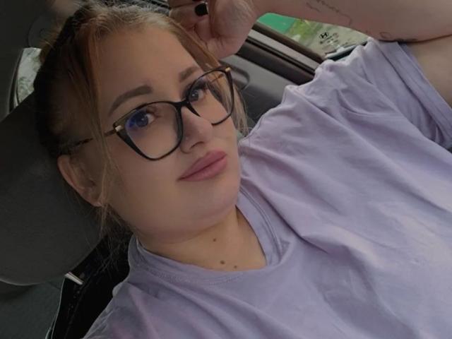 RadaRori con gli occhiali, rossetto rosa chiaro e una camicia viola, è seduta in auto, appoggiata allo schienale, mostrando un tatuaggio sul polso, con i capelli castani raccolti in un'acconciatura disordinata..