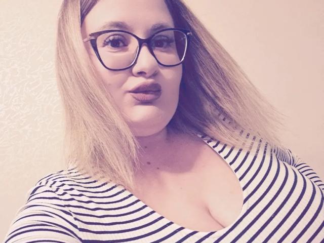 RadaRori with blonde shoulder-length hair, wearing black-rimmed glasses and a striped top with a deep V-neck, showing her big boobs. Light skin, subtle makeup, pouty lips.