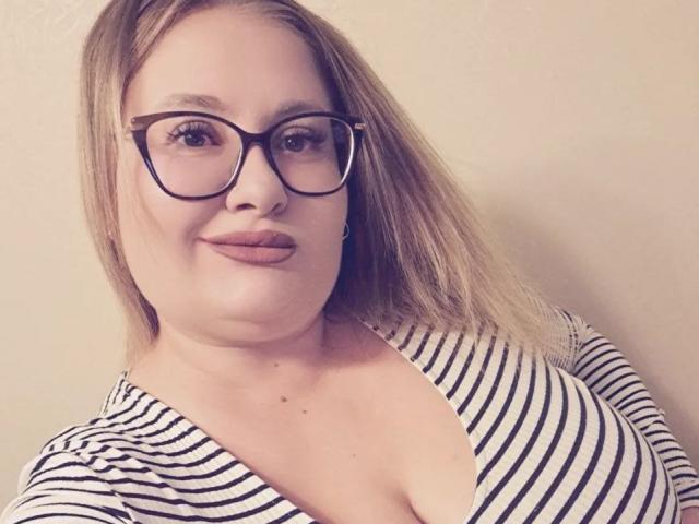 RadaRori with glasses, blonde hair, pouty lips, and a striped shirt showing her big boobs, standing against a plain wall. She's smiling softly at the camera.