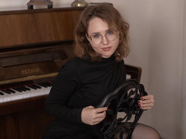 TeresaBirt with wavy brown hair, glasses, black dress, and red nails, sitting on a wooden piano bench, holding ornate black backrest, looking at camera with subtle smile.