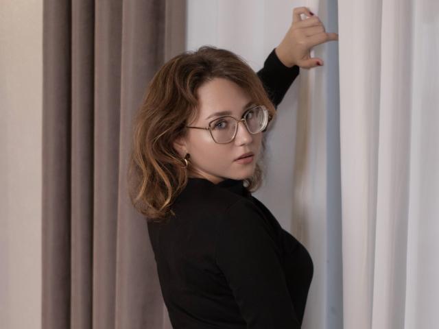 TeresaBirt with wavy brown hair, wearing glasses and a black top, looking over her shoulder, raising her right arm against white and grey curtains.