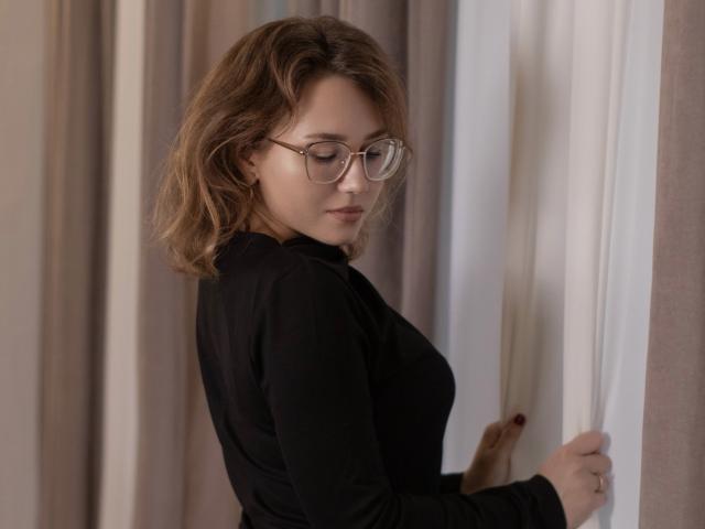 Teresa Birt con gli occhiali, seno di media grandezza, top nero, che tiene una tenda, capelli castani ondulati, che guarda in basso, luce soffusa, trucco leggero, espressione delicata..
