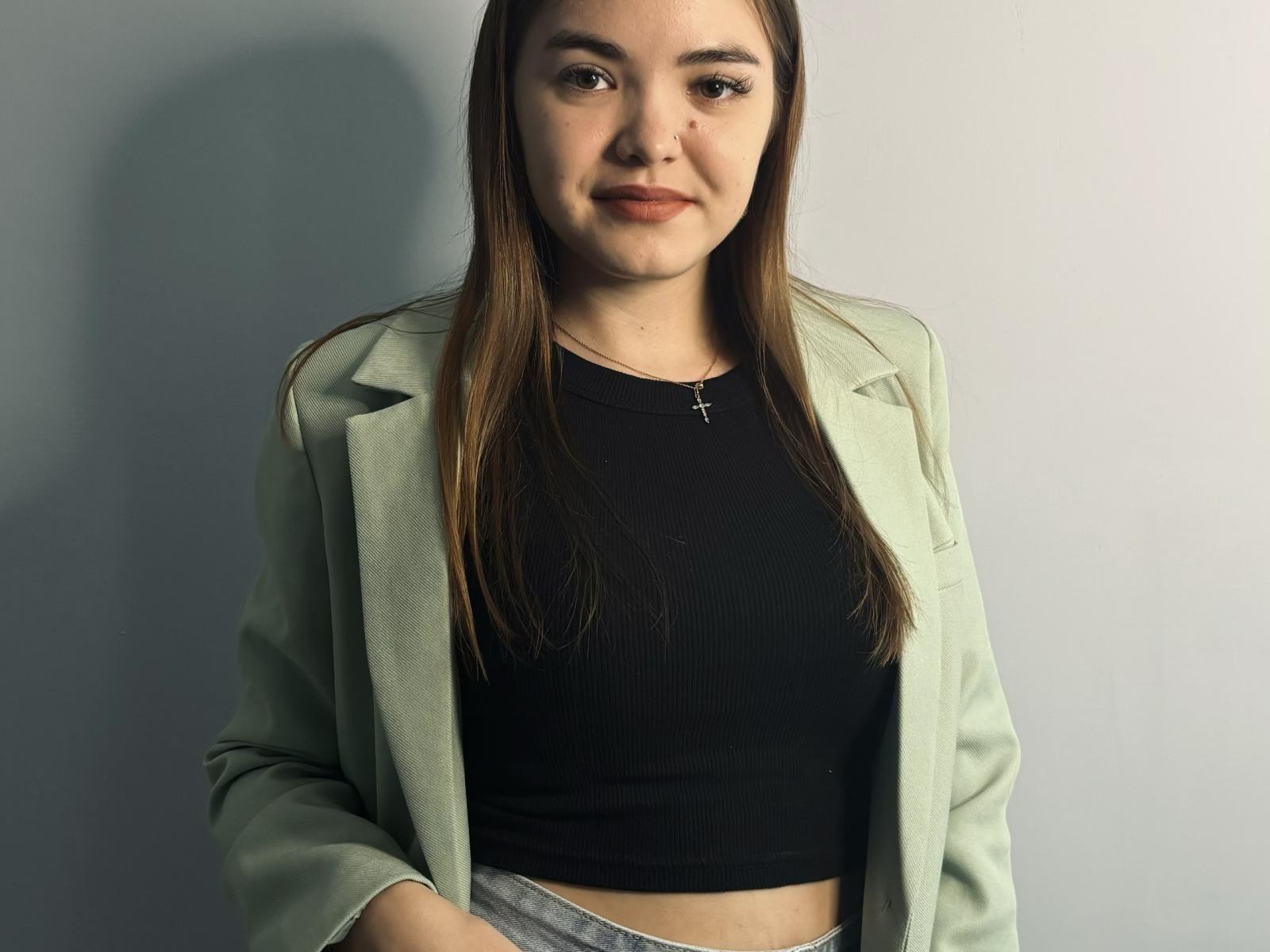 MarciPinnix with long brown hair, light green blazer, black crop top, cross necklace, and grey pants, standing against a grey wall, looking slightly to the right.