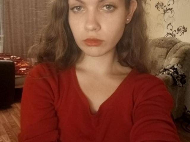 BrettRamsaroop, met golvend bruin haar, een rood shirt met lange mouwen en rode lippenstift, staart naar de camera en toont kleine borsten, een slank figuur en bleke huid, in een slecht verlichte kamer..