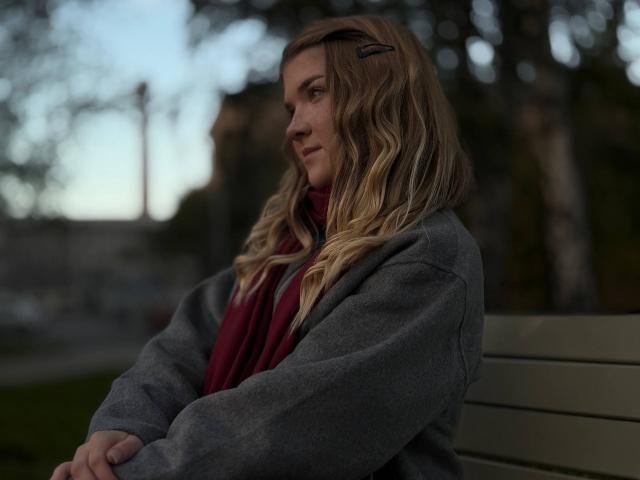 NorahHabrock with wavy blonde hair, wearing a gray jacket and red scarf, sitting on a wooden bench, arms crossed, looking to the side, hairclip in her hair.