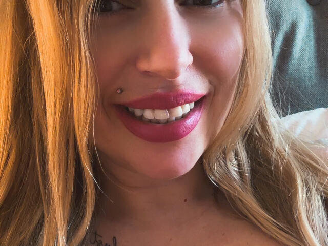 Il viso di XimenaBella, che mostra un sorriso con rossetto rosso, un piercing al naso e capelli biondi, con un tatuaggio sul collo e una camicia bianca visibile..