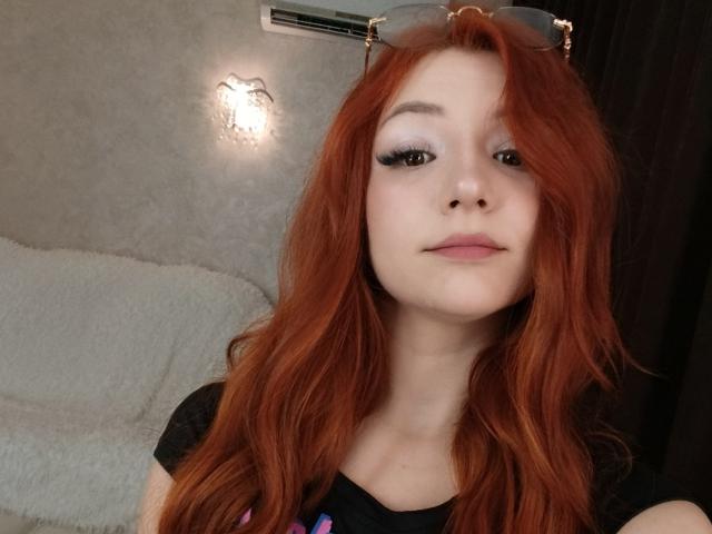 AmandaGrasy with red hair, wearing black shirt, eyeglasses on head, light makeup, and pink lips, looking at camera with a neutral expression. Sheer eyelashes, soft pink lips.