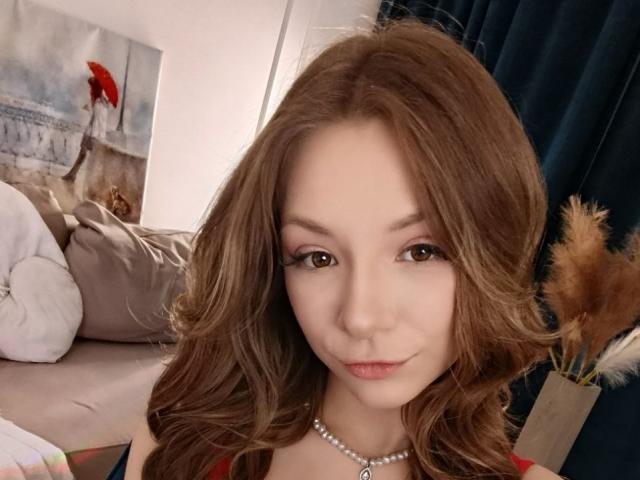 AmandaGrasy with wavy brown hair, light skin, and brown eyes, wearing a red top and pearl necklace, has small tits and a cute smile. Bedroom background, dark curtain.