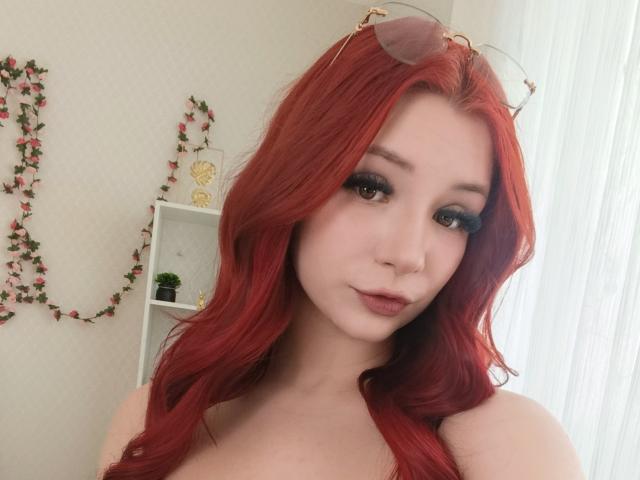 AmandaGrasy with red hair, wearing glasses on her head, has dark eyeliner, long lashes, and pink lipstick, showing her tits, with a cute, sexy expression.