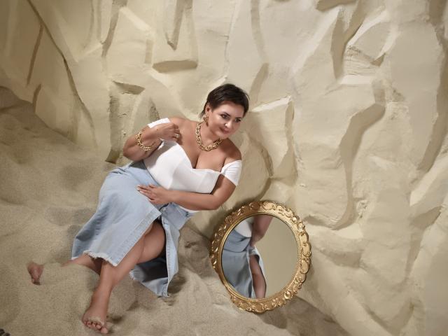 AnnaCorlane, barefoot, sitting on sandy floor, white off-shoulder top, blue denim skirt, gold necklace, bracelet, gold mirror, short brown hair, sexy, large tits, relaxed, wall behind.