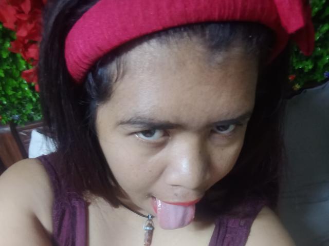 MariahSquirting69 with dark hair, red headband, and tongue out, wearing a maroon top, has a small nose ring and a pendant, looking up with a slightly annoyed expression.