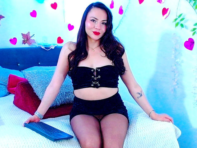 EmelySofia with dark hair, red lipstick, black strapless top, black mini-skirt, and black pantyhose, sitting on a bed with a remote, showing a tattoo on her left arm.