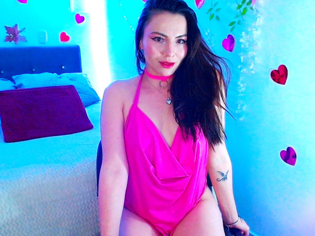 EmelySofia with long dark hair, pink halter one-piece, red choker, butterfly tattoo on left arm, sitting on bed, looking at camera, blue-lit room, pink heart stickers.