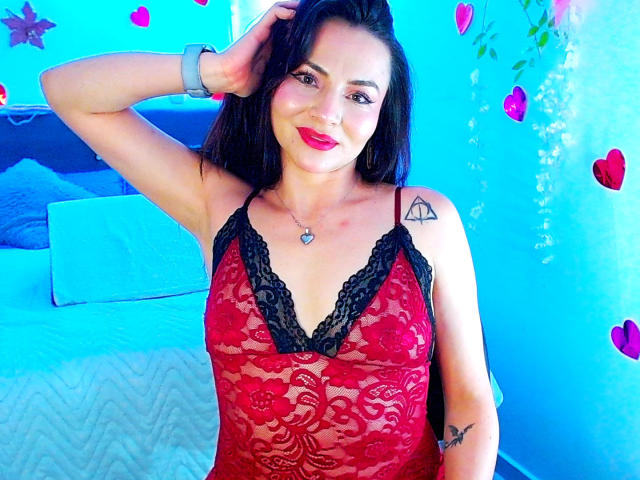 EmelySofia vestindo lingerie vermelha com detalhes em renda preta, mostrando seus seios através de um tecido transparente, sorrindo com batom vermelho, cabelo preto e uma tatuagem em formato de triângulo no ombro direito..