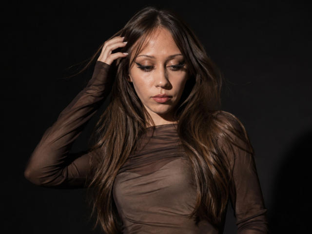 GillianSerrao with long brown hair, wearing a sheer brown top, has her hand in her hair, looking down with a serious expression, tits slightly visible through the fabric.
