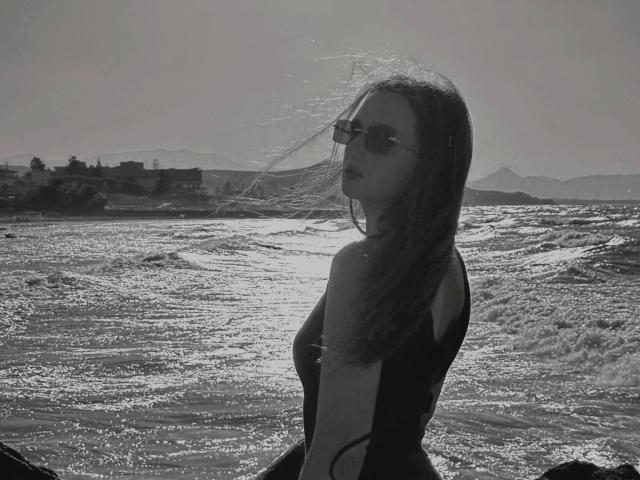 CyrenaLuxe with dark hair, wearing a black one-piece swimsuit, sunglasses, and a windblown expression, standing in shimmering water with sunlit waves. Her ass is partially visible.