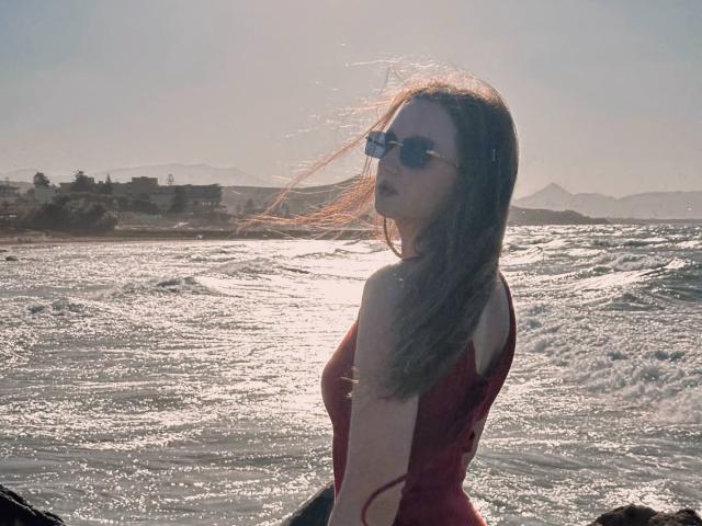 CyrenaLuxe with long brown hair, wearing a red dress and sunglasses, stands by a sunlit, wavy sea, her back slightly to the camera, looking over her shoulder.
