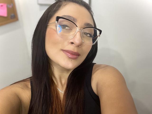 AmeliRoux has long brown hair, wearing clear glasses and a black sleeveless top, with pink lips and a subtle smile, showing her face and upper body in the photo.