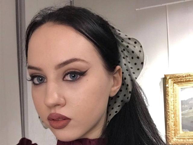 JannaCorsaro has pale skin, blue eyes with thick black eyeliner, and wears a polka-dotted headscarf, with dark red lipstick and a serious expression. Her tits are not visible.