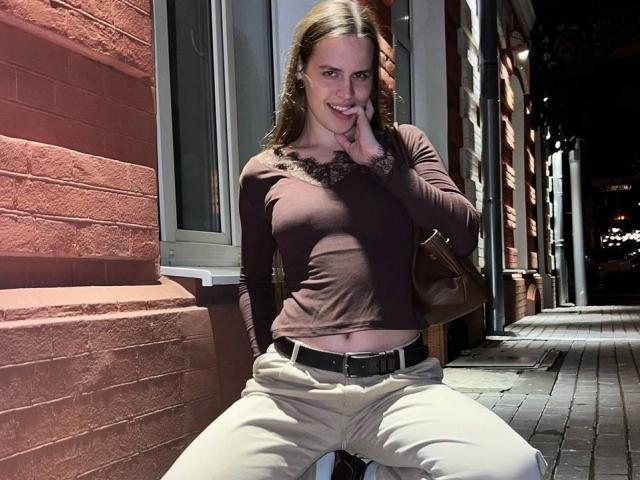 SandiWimbs with brown top, white pants, black belt, brown bag, and wavy hair, smiling, hand on cheek, sitting on white chair, brick wall, daytime, sidewalk.