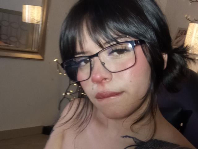 A BabeSix tem cabelo preto, usa óculos e tem um piercing no nariz. Ela está exibindo seus lábios carnudos e uma pequena tatuagem no pescoço. Seus seios estão parcialmente visíveis..