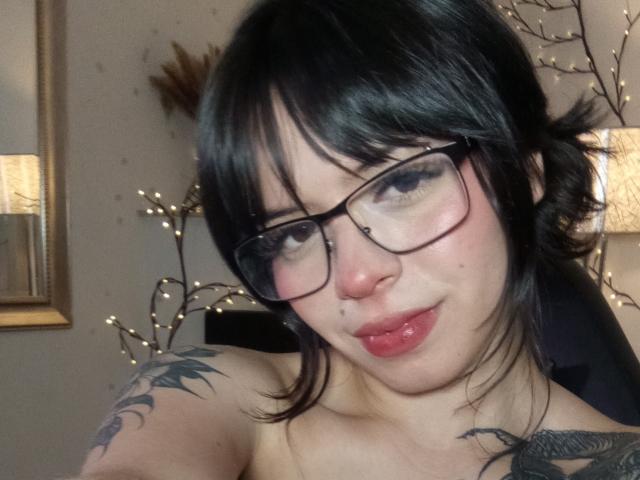 BabeSix with black hair, glasses, pink lips, nose ring, tattoos on shoulders, slight smile, soft lighting, looking at camera, semi-nude, bare shoulders, casual selfie.