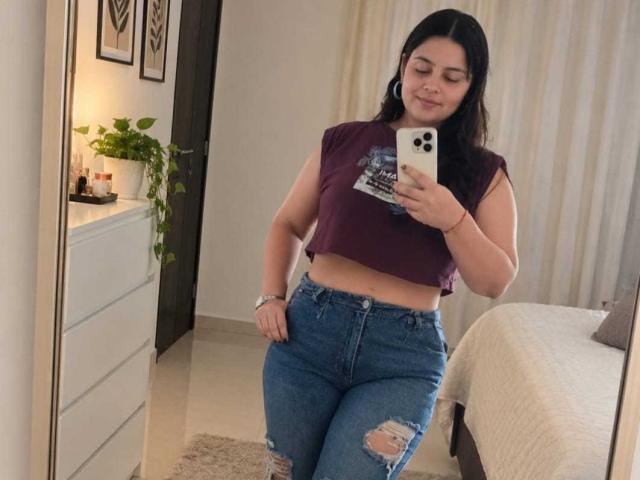 ScarletRebel with dark hair, medium tits, wearing a purple crop top and ripped blue jeans, taking a mirror selfie in a bedroom, smiling slightly.