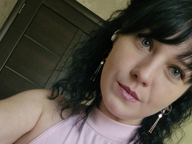 WenTina has curly black hair, green eyes, and pink lipstick. She's wearing a pink sleeveless top and gold earrings, smiling with glossy pink lips. Her ass is not visible.