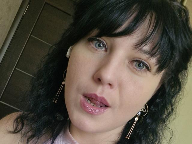 WenTina has black wavy hair, blue eyes, and pink glossy lips with slightly parted teeth. She's wearing pink earrings and a pink top, looking directly at the camera.