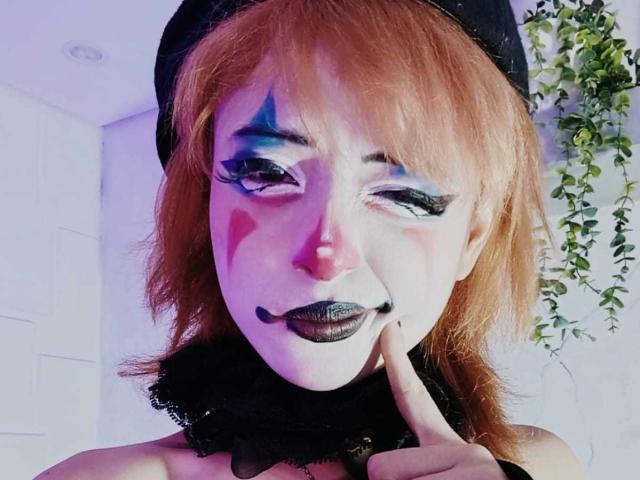 ViolettClay, con i capelli rossi, il rossetto nero, il trucco sul viso in colori blu e rosa e l'eyeliner nero, che tocca il mento con un dito, indossa un cappello nero e un colletto di pizzo..