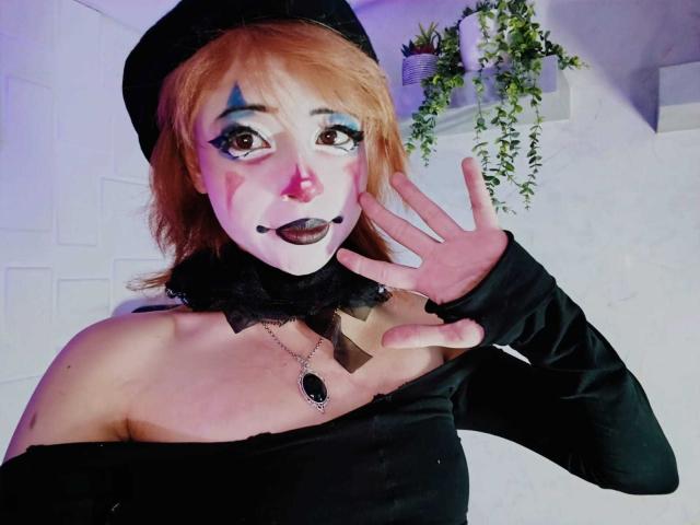ViolettClay hat rotes Haar, schwarzen Lippenstift und weiße Gesichtsbemalung mit blauen Akzenten. Sie trägt einen schwarzen Hut und ein Top mit einem Schulterteil, der herunterfällt, und hebt ihre Hand, deren Nägel schwarz lackiert sind..