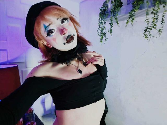 ViolettClay, con piel pálida, cabello naranja, una blusa negra con un hombro descubierto, un collar negro, maquillaje azul en los ojos, lápiz labial negro y un sombrero negro, sosteniendo su clavícula, en un estilo gótico..