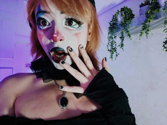 ViolettClay with pale skin, orange hair, and black lipstick, hand over mouth, black nails, black off-shoulder top, necklace, and purple-blue eye makeup, in a dimly lit room.