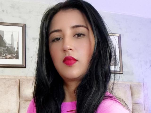 NicollJeans has black hair, pink lipstick, and brown eyes, wearing a pink top, with a neutral expression. Her face is centered, showing a slightly parted, pink-lipsticked mouth.