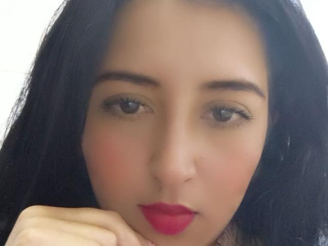 NicollJeans has a cute face with dark hair, brown eyes, and red lipstick. She's resting her chin on her hand, looking slightly bored. Her tits are not visible.