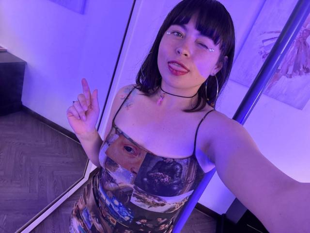 Iniridax, with dark brown hair and bangs, winking, wearing a colorful eye-pattern dress, holding up a finger, has tattoos and a choker, in a purple-lit room.
