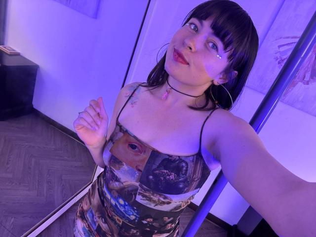 Iniridax with dark brown hair and blue eyes, wearing a colorful dress with facial prints, has a pink gem necklace, hoop earrings, and a tear on her cheek, taking a selfie.