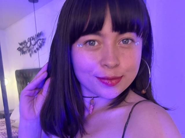 Iniridax with dark brown hair, blue eyes, and sparkling eye makeup, wearing red lipstick, hoop earrings, and a pink gem necklace, smiling in purple-lit room.