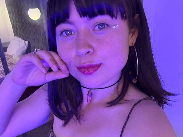 Iniridax with purple-blue lit face, dark brown hair, bangs, small crystal teardrop on cheek, red lipstick, black choker, pendant, hoop earrings, hand touching cheek, smiling.