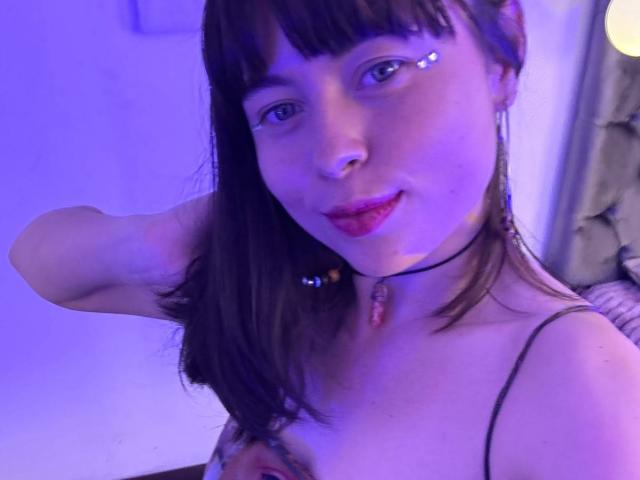Iniridax with dark brown hair, blue eyes, and nose ring, smiling, wearing a choker with a gem, and showing small tits with a hand on her chest, under purple light.