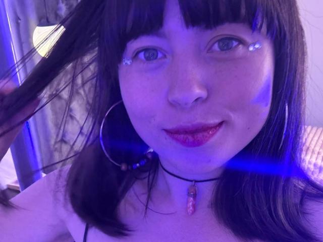 Iniridax with purple-blue light on face, holding black hair, wearing red lipstick, hoop earrings, choker with pink stone, blue light flares, smiling, dark straight hair, fair skin.