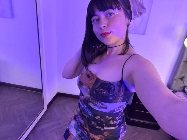 Iniridax with short black hair, wearing a colorful spaghetti-strap top, purple-blue room lighting, black choker, and subtle eye makeup, holding a selfie. Boobs visible, cute expression.