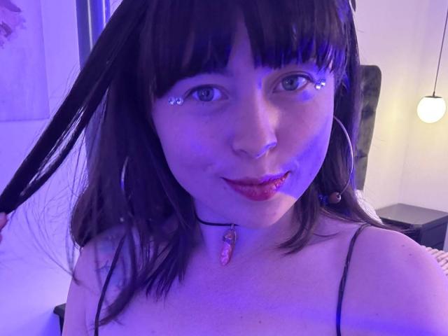 Iniridax has purple-blue lit face, dark brown hair, bangs, and is holding her hair with a tongue out, wearing hoop earrings and a choker with a pink gem.