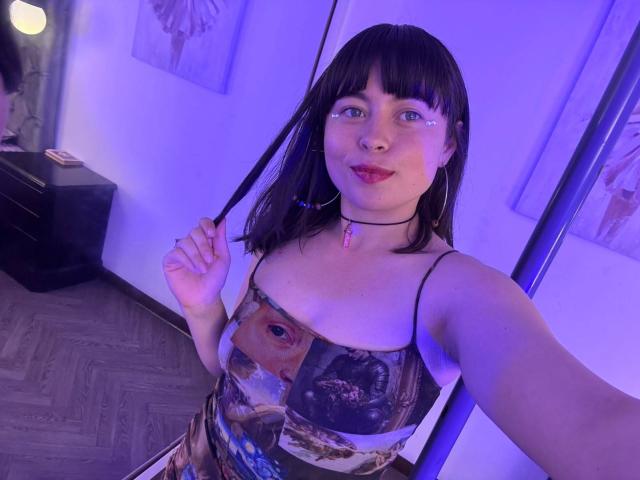 Iniridax with black hair, blue eyes, wearing a colorful, eye-patterned dress, holding a strand of hair, purple light, hoop earrings, black choker, and pink gem necklace, smiling.